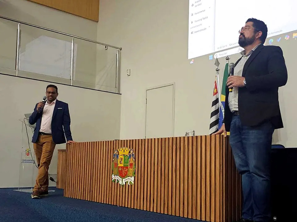 Paulo Moraes e Darniton Viana, os instrutores do curso, falando ao público em um auditório, com o sistema projetado ao fundo